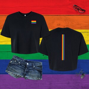 Pu&ograve; includere: Una t-shirt nera corta con una piccola bandiera arcobaleno sul petto a sinistra e una striscia verticale arcobaleno centrata sul retro. La maglietta &egrave; mostrata insieme a un paio di pantaloncini di jeans blu con orlo sfrangiato.