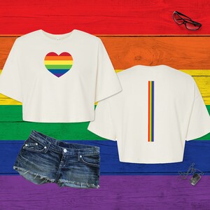 Pu&ograve; includere: Una t-shirt corta bianca con un cuore color arcobaleno sul davanti e una striscia verticale arcobaleno sul retro, abbinata a pantaloncini di jeans blu, occhiali con montatura nera e un ciondolo a piastrina argentato su una catenina.