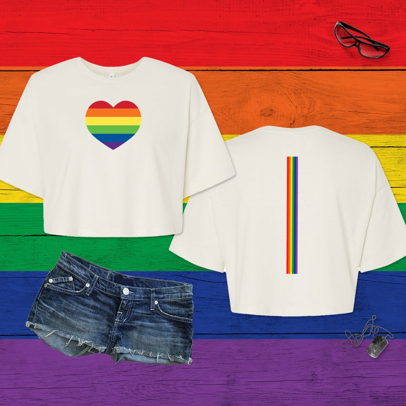 Pu&ograve; includere: Una t-shirt corta bianca con un cuore color arcobaleno sul davanti e una striscia verticale arcobaleno sul retro, abbinata a pantaloncini di jeans blu, occhiali con montatura nera e un ciondolo a piastrina argentato su una catenina.