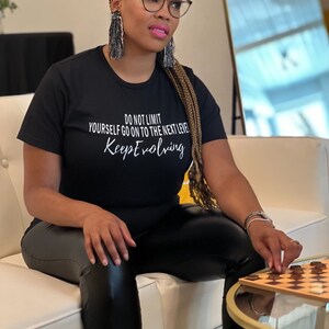 Puede incluir: Una mujer con una camiseta negra con el texto "Do not limit yourself go on to the next level Keep Evolving" en letras blancas. Está sentada en un sofá blanco y jugando una partida de damas en un tablero de madera.