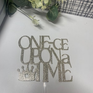 May include: Silver glitter cake topper with the words "ONE-CE UPON a TIME" and a crown. The topper is attached to a clear plastic stick, ideal for a birthday or special occasion.