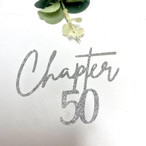 May include: A silver glitter cake topper featuring the text Chapter 50 in a cursive and sans-serif font combination. The topper is positioned below a small sprig of green eucalyptus leaves, creating a decorative accent for a milestone celebration.