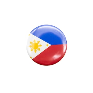 May include: A round button pin featuring the flag of the Philippines. The design includes a blue upper section, a red lower section, and a white triangle with a yellow sun and stars. The pin is set against a white background.
