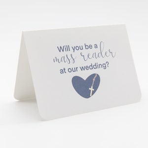 以下が含まれることがあります： 結婚式でミサの読者になってくれませんか？と書いてある青い文字の白いカード。青いハートの中に金の十字架が入っています。