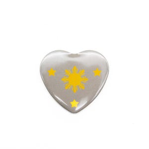 May include: A heart-shaped pin with a gray background and a yellow sun with a starburst design and three yellow stars.