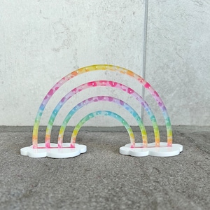 May include: A rainbow-colored acrylic earring holder shaped like a cloud with three rainbow arches. The arches are decorated with a white polka dot pattern.