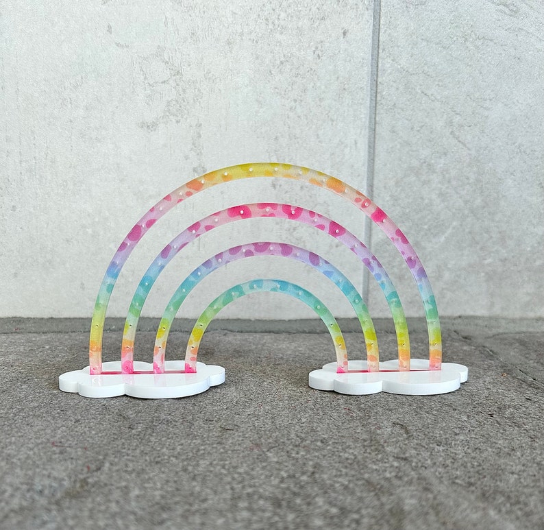 May include: A rainbow-colored acrylic earring holder shaped like a cloud with three rainbow arches. The arches are decorated with a white polka dot pattern.