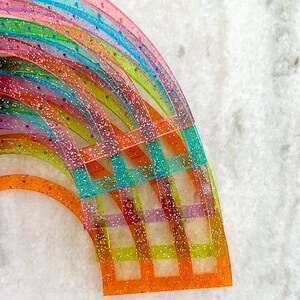 Pu&ograve; includere: Un set di righelli in plastica arcobaleno con glitter incorporati nella plastica. I righelli sono disposti a forma di semicerchio, con il righello pi&ugrave; grande in basso e il pi&ugrave; piccolo in alto.