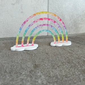 May include: A rainbow-colored, multi-layered, plastic rainbow with a white cloud base. The rainbow is decorated with small pink stars.