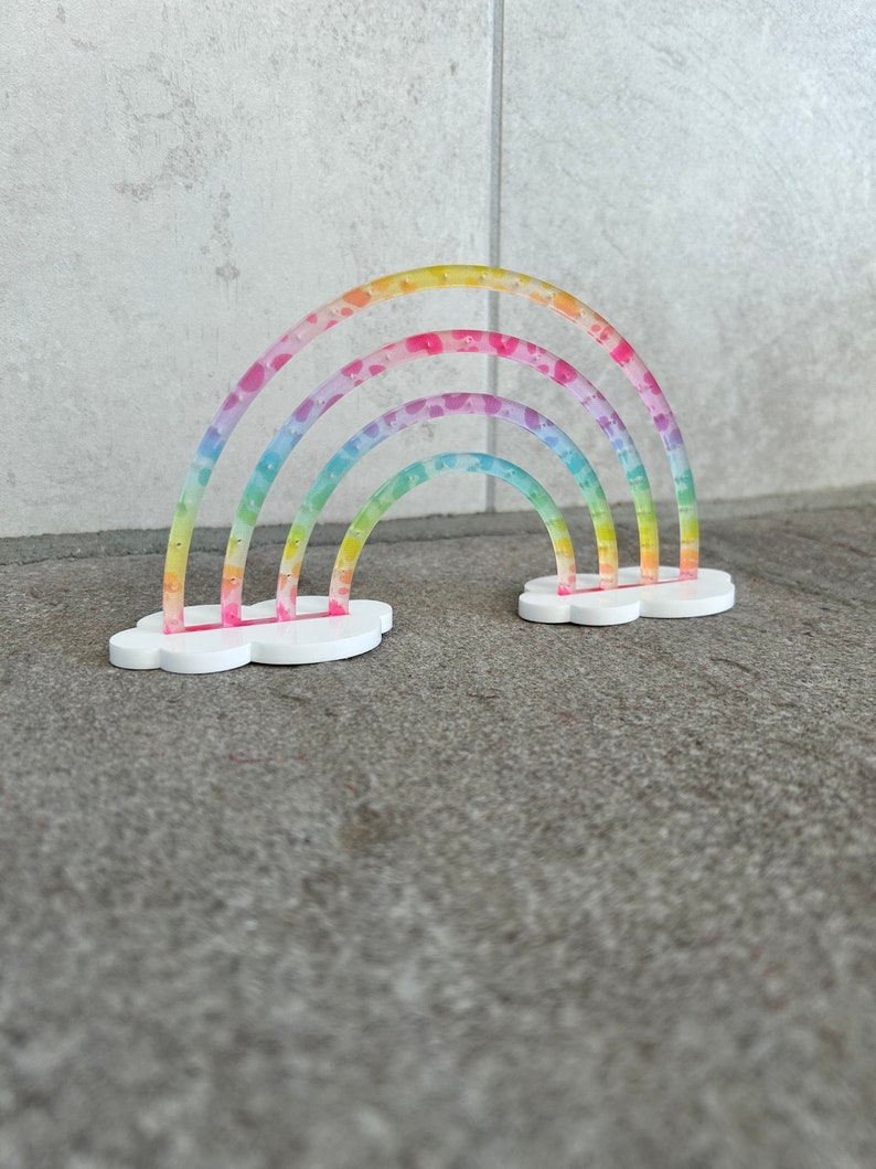May include: A rainbow-colored, multi-layered, plastic rainbow with a white cloud base. The rainbow is decorated with small pink stars.