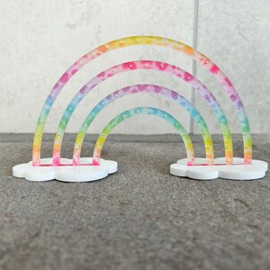May include: A rainbow-colored earring holder with a cloud base. The holder has three rainbow arches with small holes for earrings.