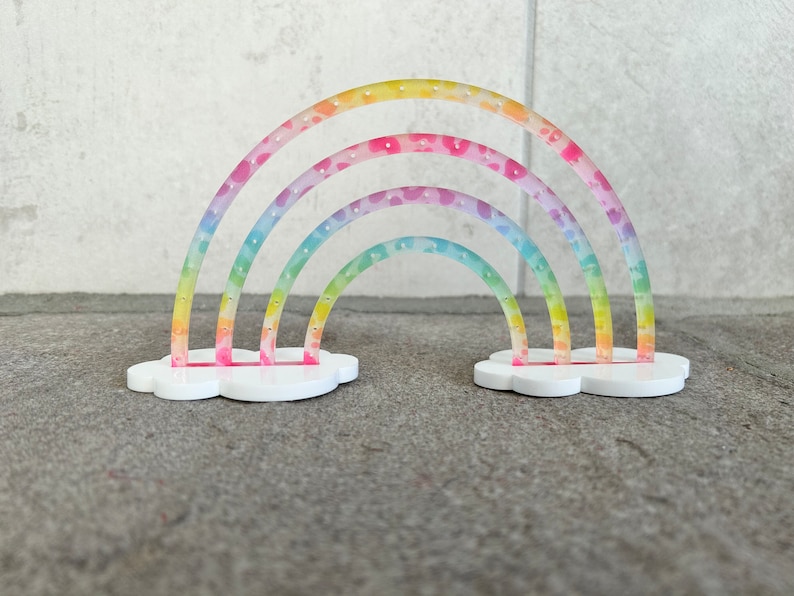 May include: A rainbow-colored earring holder with a cloud base. The holder has three rainbow arches with small holes for earrings.