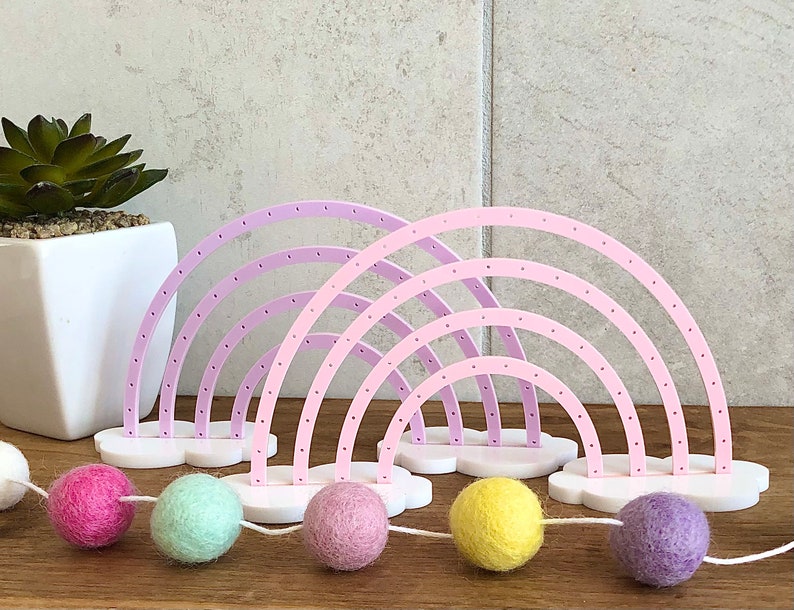 Pu&ograve; includere: Un porta orecchini a forma di arcobaleno rosa e viola con una base a forma di nuvola. Il porta orecchini ha pi&ugrave; file di fori per gli orecchini. In primo piano c'&egrave; una corda di palline di feltro colorate.