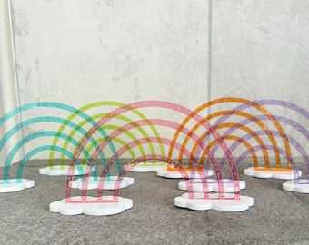 May include: A set of seven rainbow-colored plastic arches, each with a cloud base. The arches are arranged in a row, with the largest arch in the back and the smallest arch in the front. The arches are made of clear plastic with a glittery finish.