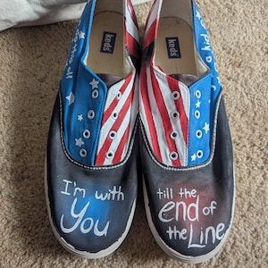 Puede incluir: Un par de zapatos de lona negros pintados con un diseño de bandera estadounidense. Los zapatos tienen las palabras "I'm with You" y "Till the end of the line" escritas en ellos.