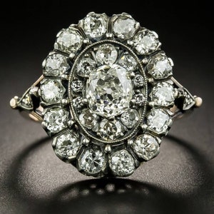 Puede incluir: Un anillo de diamantes vintage con una gran piedra central redonda rodeada de diamantes más pequeños. El anillo está engastado en plata y tiene un delicado diseño floral.