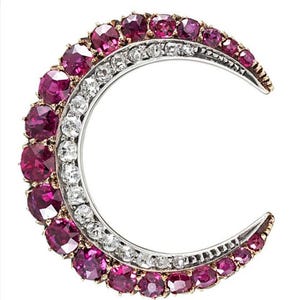 Puede incluir: Un broche con forma de media luna con una fila de piedras de color rojo rubí y una fila de diamantes transparentes.