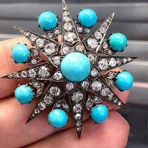 Puede incluir: Un broche de estrella de plata con un gran cabujón turquesa en el centro y cabujones turquesa más pequeños alrededor de los bordes. El broche está engastado con numerosos diamantes pequeños.