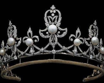 Puede incluir: Una tiara de plata con un delicado diseño floral y una fila de perlas. La tiara está sobre un fondo negro.