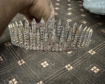 Könnte beinhalten: Ein silberfarbenes Diadem mit einem goldfarbenen Band. Das Diadem hat eine Reihe von vertikalen, spitzen Elementen, die jeweils mit mehreren klaren, funkelnden Steinen verziert sind. Die Basis des Diadems ist mit quadratischen Steinen besetzt.