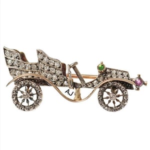 May include: A silver and gold vintage car brooch with diamond accents and green and pink gemstones. The brooch is in the shape of a classic car with two seats and a steering wheel. The car is detailed with a diamond-encrusted body and wheels. The brooch is signed "AJ".