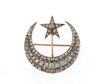 May include: A vintage brooch featuring a crescent moon and star design. The brooch is adorned with numerous small, sparkling gemstones, likely diamonds, set in a silver or platinum metal. The star is positioned above the moon, connected by a gold-toned bar.
