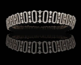 Könnte beinhalten: Ein silbernes Armband mit Diamanten und einem sich wiederholenden Muster aus ineinandergreifenden Kreisen. Das Armband spiegelt sich auf einer schwarzen Oberfläche.