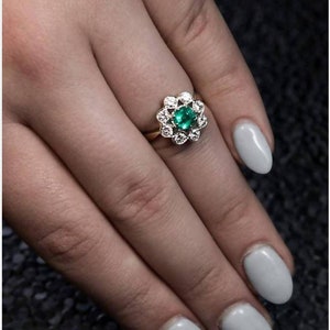 May include: A gold ring with a green emerald gemstone surrounded by smaller diamonds. The ring is on a finger with light gray nail polish.