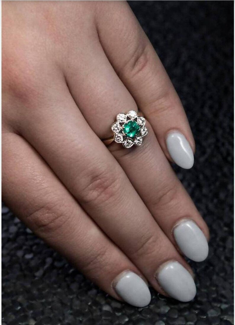May include: A gold ring with a green emerald gemstone surrounded by smaller diamonds. The ring is on a finger with light gray nail polish.