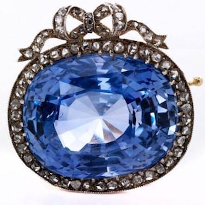 Op de afbeelding: Een grote blauwe saffier gezet in een zilveren broche met een diamanten strik. De broche is omgeven door een halo van kleinere diamanten.