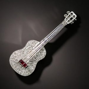 以下が含まれることがあります： 銀色のギター型のブローチ。ブローチの本体には、小さな輝く石がちりばめられています。長方形の赤い石が弦の付け根近くにセットされています。ブローチは暗い背景にセットされています。