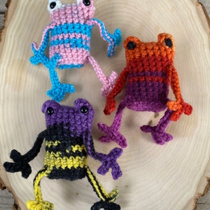 May include: Three crocheted frog toys. One is pink and blue, one is orange and purple, and one is purple and yellow.