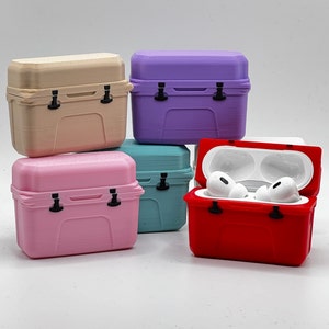 May include: A collection of colorful, miniature cooler-shaped cases for wireless earbuds. The cases are in various pastel colors, including pink, teal, purple, and beige. One red case is open, revealing white earbuds inside. Each case has a black latch.