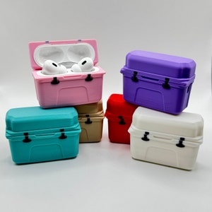 May include: A collection of colorful, miniature cooler-shaped cases for wireless earbuds. The cases are in various colors, including pink, teal, purple, red, white, and tan. One pink case is open, revealing white earbuds. Each case has a latch and a handle.