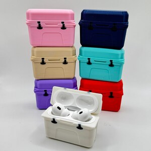 May include: A stack of colorful miniature cooler-shaped cases. The cases are in various colors, including pink, beige, purple, blue, turquoise, and red. One white case is open, revealing a pair of white earbuds. The cases have black latches.