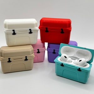 May include: A collection of colorful, miniature cooler-shaped cases for wireless earbuds. The cases are in various colors, including white, red, pink, purple, tan, and teal. One teal case is open, displaying white earbuds.