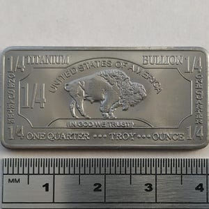 May include: A silver bullion bar with the inscription "1/4 Titanium Bullion 1/4 oz. Gold .9999 Fine" and "United States of America" with a bison image and the text "In God We Trust" and "1/4 One Quarter...Troy...Ounce 1/4".