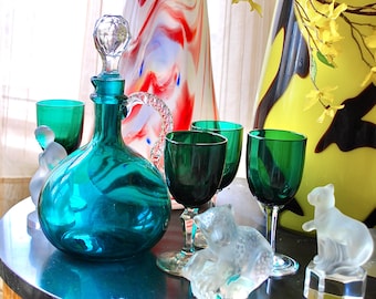 May include: A teal glass decanter with a clear glass stopper and four teal glass wine glasses on a table with two clear glass figurines.