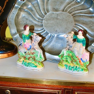 Könnte beinhalten: Zwei antike Porzellanfiguren. Jede Figur zeigt eine Person, die auf einem Hirsch reitet, mit farbenfrohen Details, darunter grüne, rosa und blaue Akzente. Die Figuren sind vor einem dekorativen Metallteller platziert.