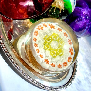 May include: A white glass paperweight with a swirling pattern of orange and green flowers on a silver tray.