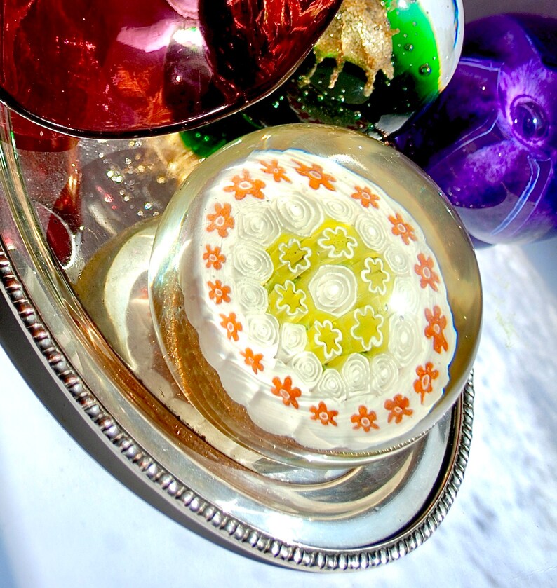 May include: A white glass paperweight with a swirling pattern of orange and green flowers on a silver tray.