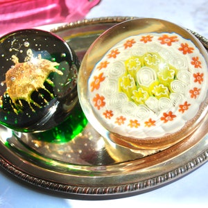 May include: Two glass paperweights on a silver tray. One paperweight is green and has a gold-colored design inside. The other paperweight is clear and has a white and orange floral design inside.