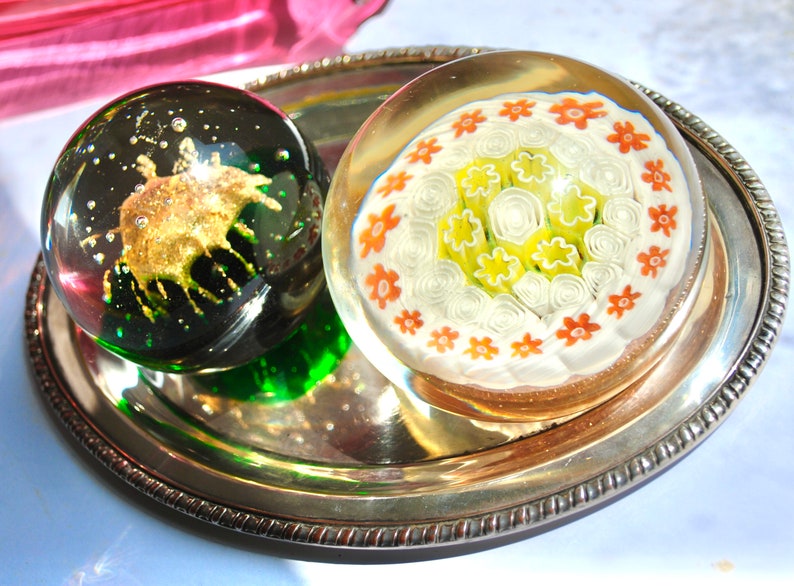May include: Two glass paperweights on a silver tray. One paperweight is green and has a gold-colored design inside. The other paperweight is clear and has a white and orange floral design inside.