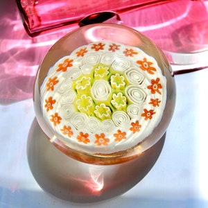 May include: A clear glass paperweight with a white, green, and orange floral design. The design features a central cluster of white and green swirls surrounded by a ring of orange flowers.