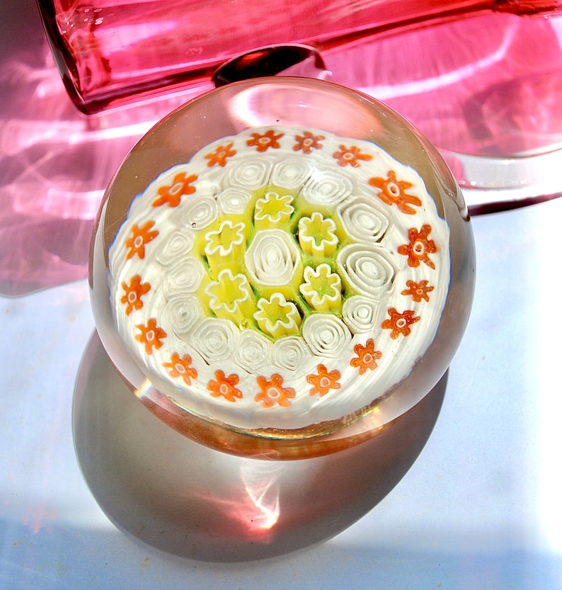 May include: A clear glass paperweight with a white, green, and orange floral design. The design features a central cluster of white and green swirls surrounded by a ring of orange flowers.
