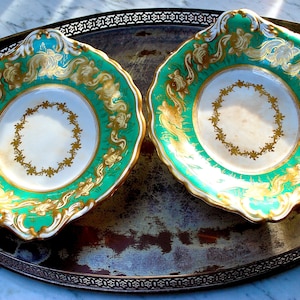 Puede incluir: Dos platos de servir de porcelana blanca adornados con detalles dorados y verdes. Los platos tienen un borde festoneado y están decorados con un patrón floral. Los platos están sobre una bandeja de plata.