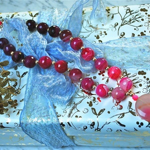 May include: A pink and purple agate bead necklace with a large pink agate pendant on a white gift box with a blue ribbon.