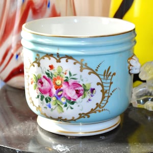 Könnte beinhalten: Ein hellblauer Porzellan-Pflanztopf mit goldenen Verzierungen und Blumendesigns. Der Topf hat zwei Löwenkopf-Griffe und einen weißen Sockel.