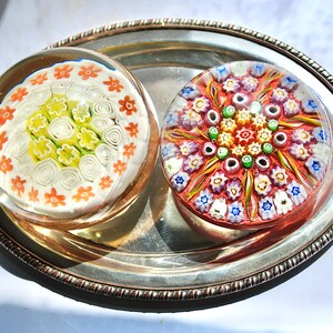 May include: Two colorful glass paperweights with intricate floral designs. One paperweight features a white background with orange and yellow flowers, while the other has a red background with multi-colored flowers.