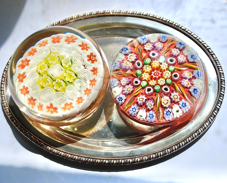 May include: Two colorful glass paperweights with intricate floral designs. One paperweight features a white background with orange and yellow flowers, while the other has a red background with multi-colored flowers.